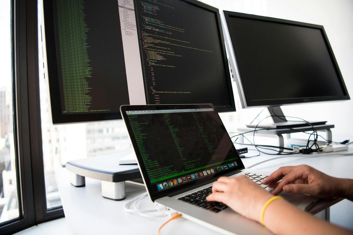 A developer writes code on a laptop in front of multiple monitors in an office setting